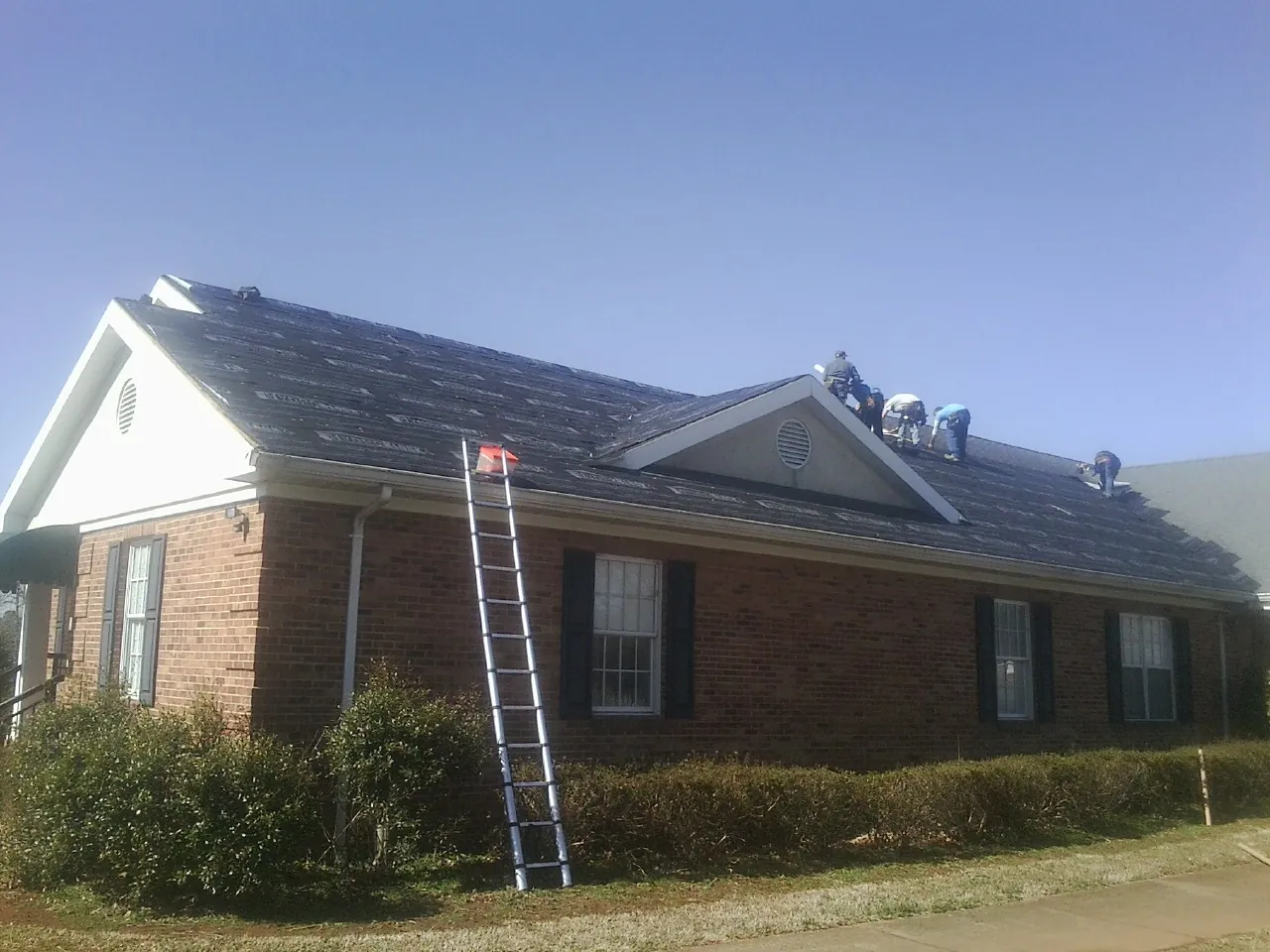 Crew tearing shingles off a brick ranch roof with a ladder set — Emergency Roof Repair work by our crew in Sunapee