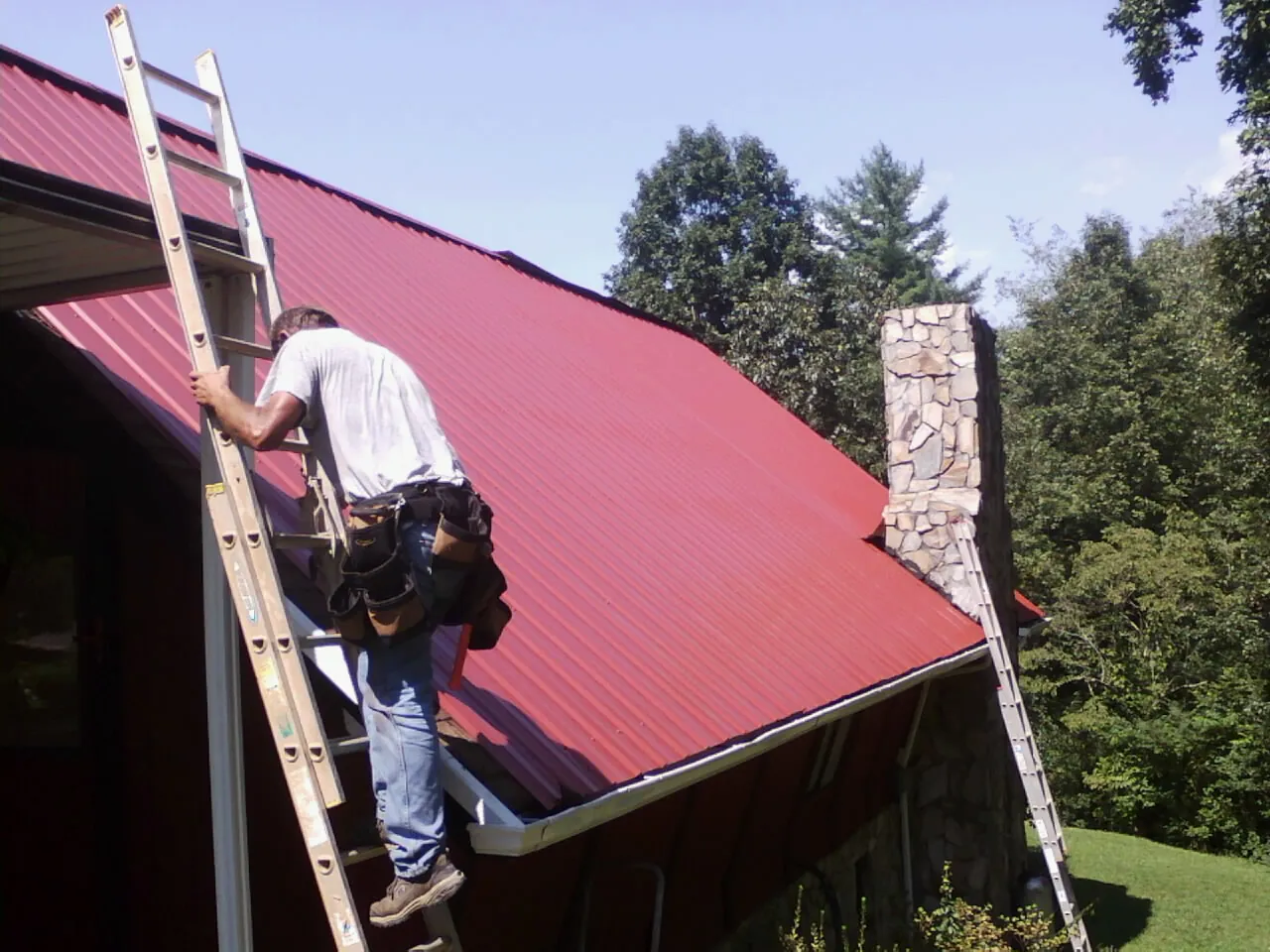Roofer on a ladder beside a red metal roof and stone chimney — Soffit & Fascia Repair by Roofing Brothers in Sunapee
